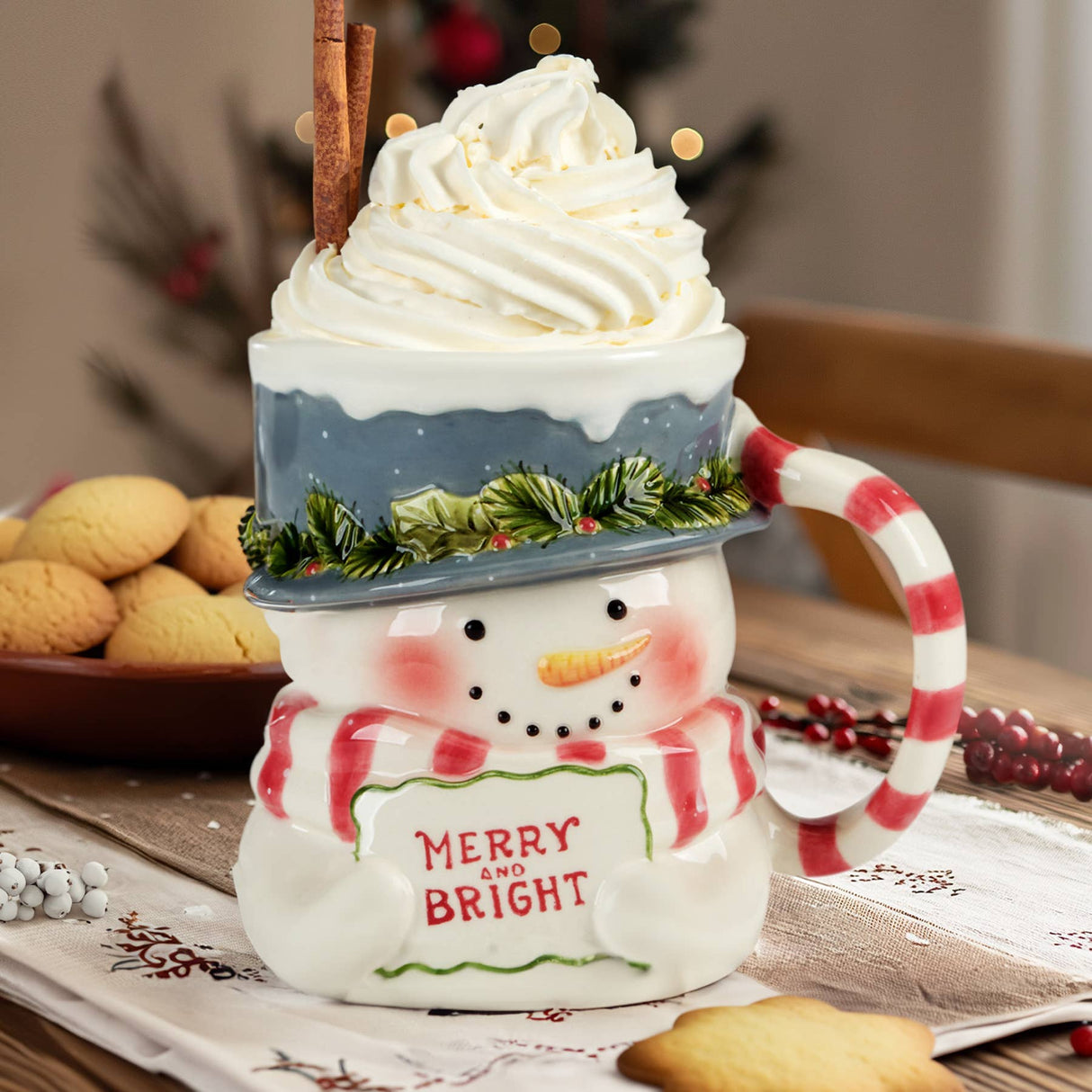 Snowman-themed mug with whipped cream and a cinnamon stick on a festive table.