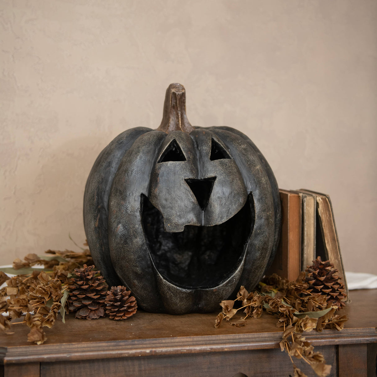 Decorative pumpkin with carved face on a wooden surface with books and pinecones.