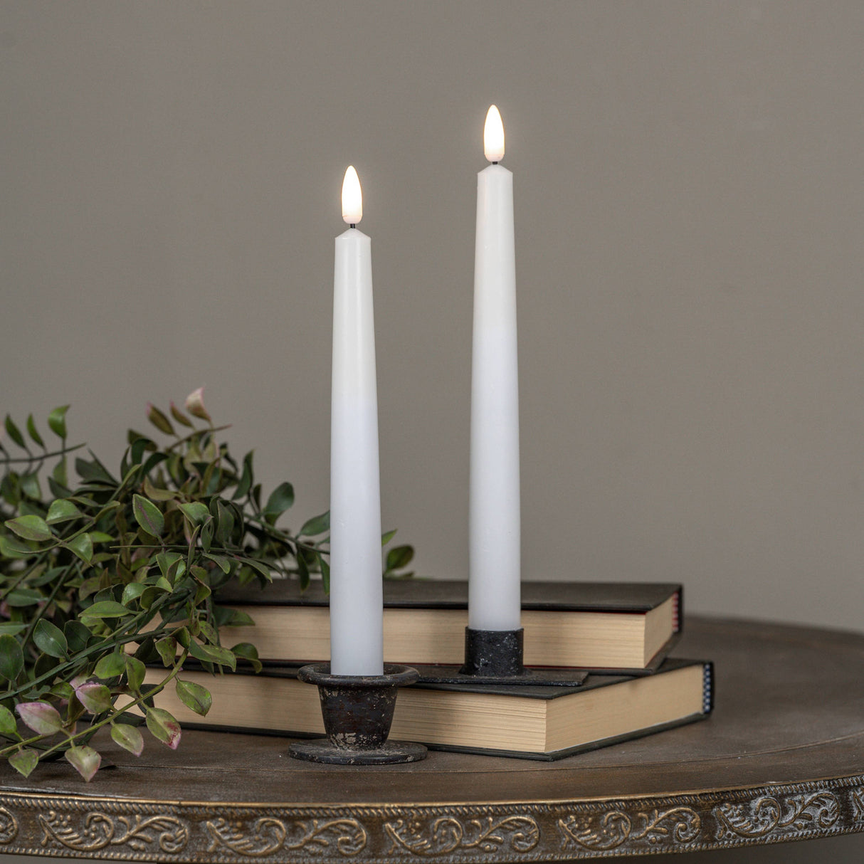 Two lit flameless candles in a decorative holder on a wooden surface with books and greenery.