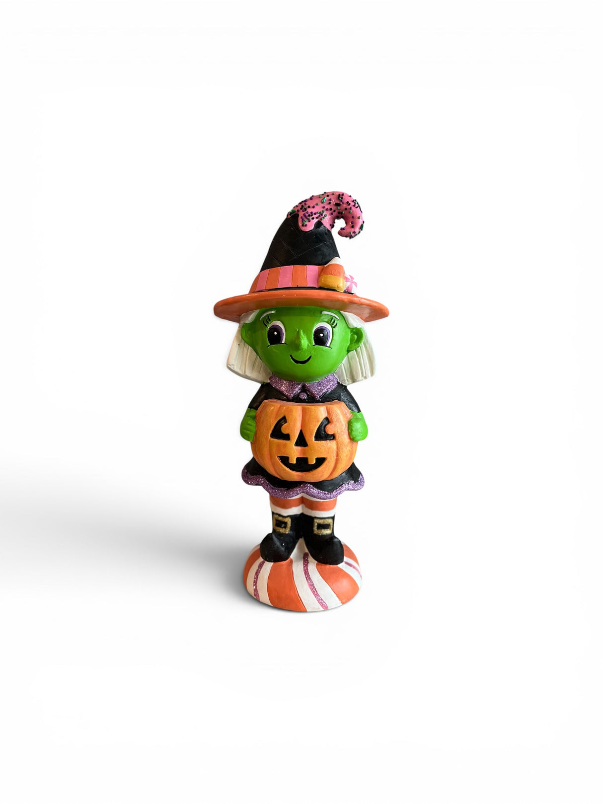Whimsical Halloween witch figurine holding a jack‑o’‑lantern, wearing a black dress with purple glitter trim, candy‑accented witch hat, striped stockings, and standing on an orange‑and‑white striped base.
