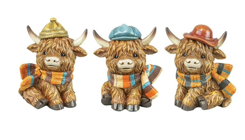 Three decorative Highland cow figurines wearing hats and scarves on a white background