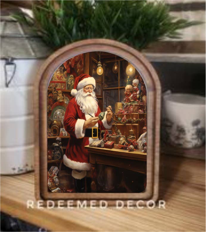 Decorative scene with Santa Claus in a workshop, framed by a wooden border, on a table with plants and a mug.