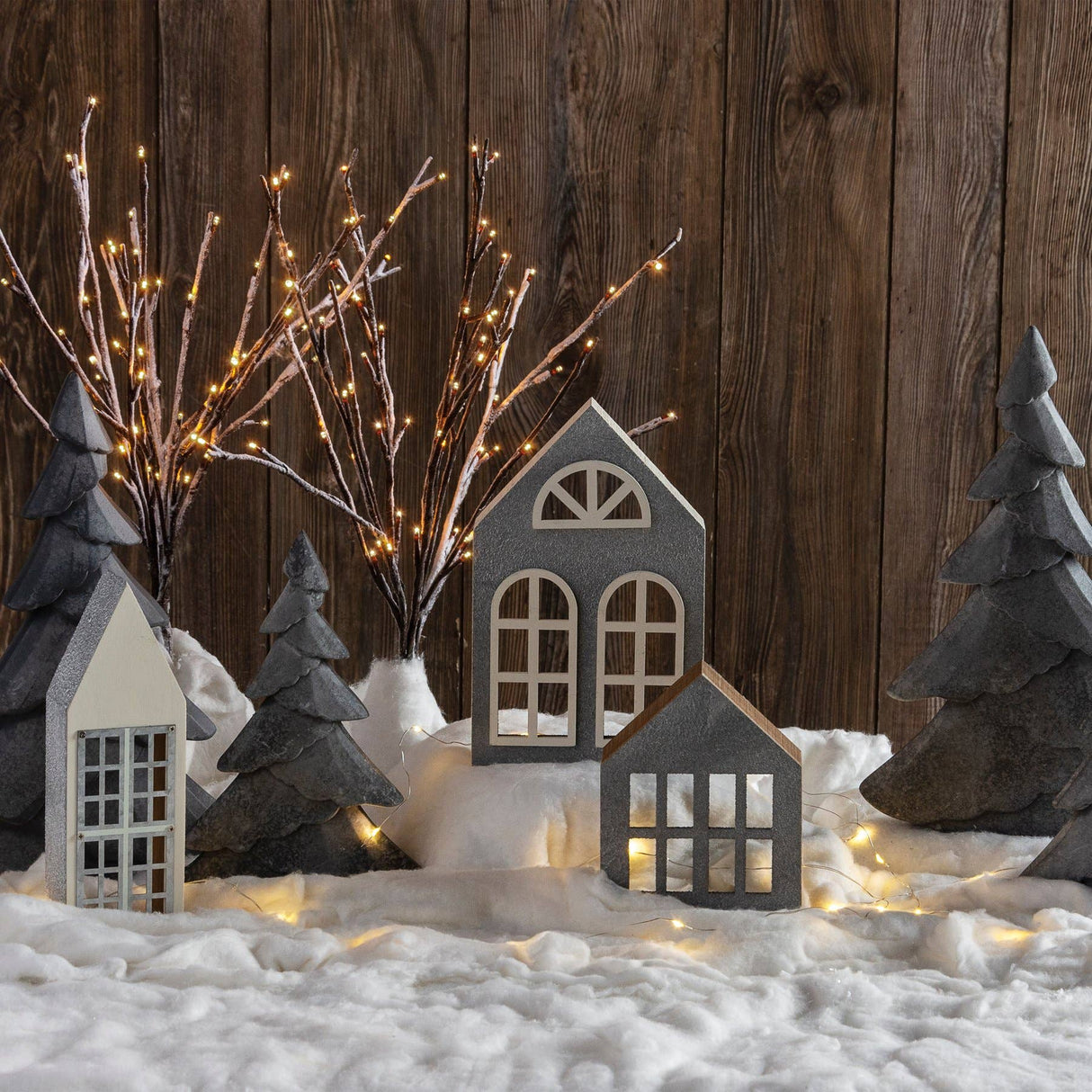 Cozy winter village display with miniature houses, evergreen trees, warm string lights, and a soft layer of artificial snow creating a festive holiday atmosphere.