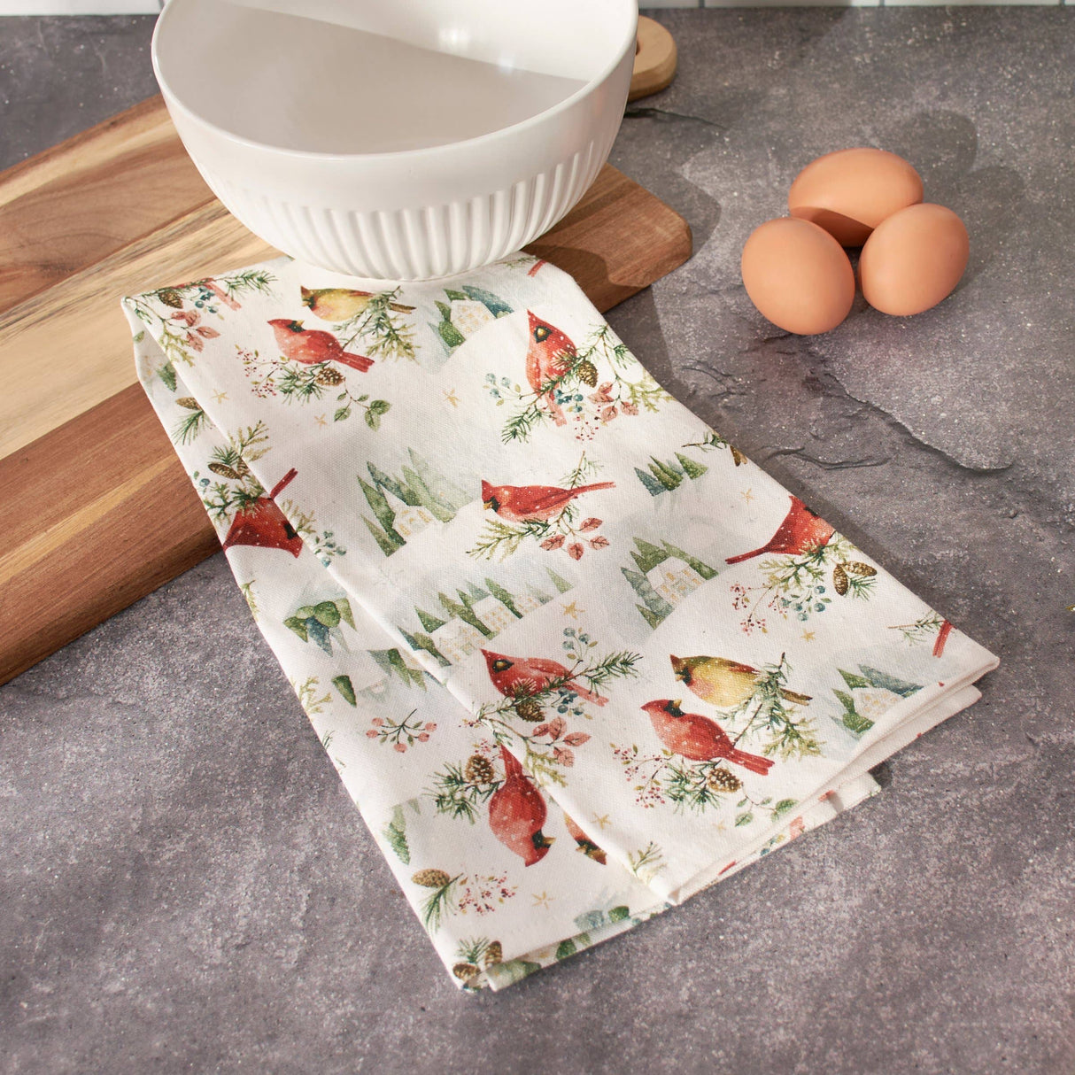 A folded kitchen towel with a red cardinal and green pine tree pattern on a cream background, styled on a gray stone surface next to a white ribbed bowl, a wooden cutting board, and three brown eggs.