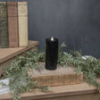 Black pillar candle styled on stacked vintage books with soft greenery, showing a realistic moving flame wick and matte black finish.