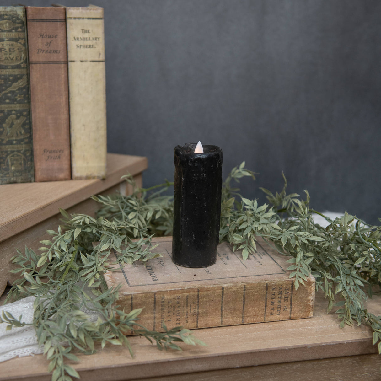 Black pillar candle styled on stacked vintage books with soft greenery, showing a realistic moving flame wick and matte black finish.