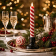 Red‑and‑white candy cane striped LED taper candle glowing in a black holder on a festive holiday table with greenery, berries, and champagne glasses in the background.