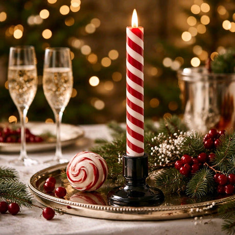 Red‑and‑white candy cane striped LED taper candle glowing in a black holder on a festive holiday table with greenery, berries, and champagne glasses in the background.