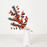 A Halloween spray with soft black pine branches and glittered orange accents arranged in a white pitcher.