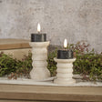 Set of two black 3D‑flame tealight candles styled on ribbed cream holders with soft greenery.