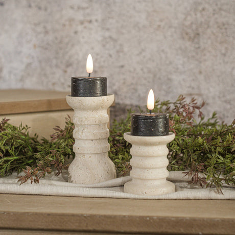 Set of two black 3D‑flame tealight candles styled on ribbed cream holders with soft greenery.