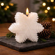 White snowflake‑shaped candle glowing on a dark stone surface with pine branches and pinecones in a cozy holiday setting.