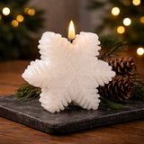 White snowflake‑shaped candle glowing on a dark stone surface with pine branches and pinecones in a cozy holiday setting.