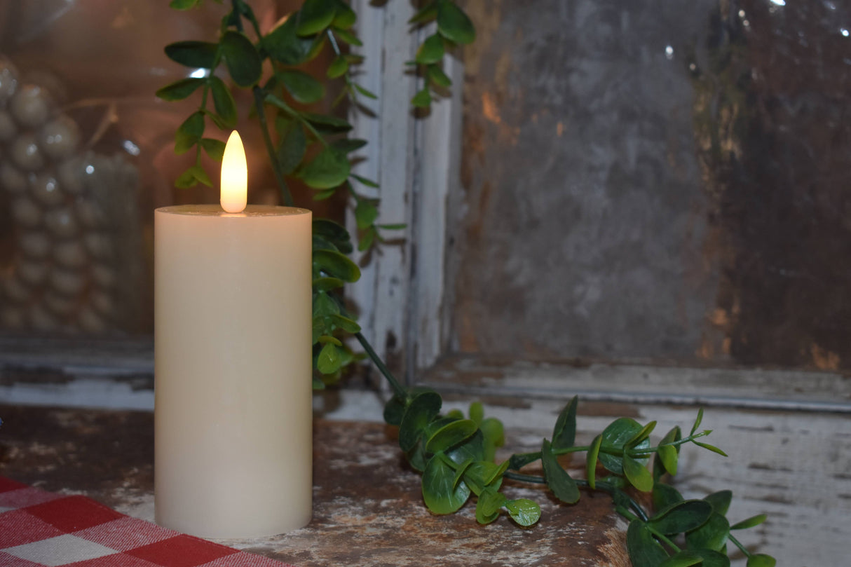 Cream LED pillar candle glowing beside a window, surrounded by a trailing green vine and rustic décor for a warm, cozy ambiance.