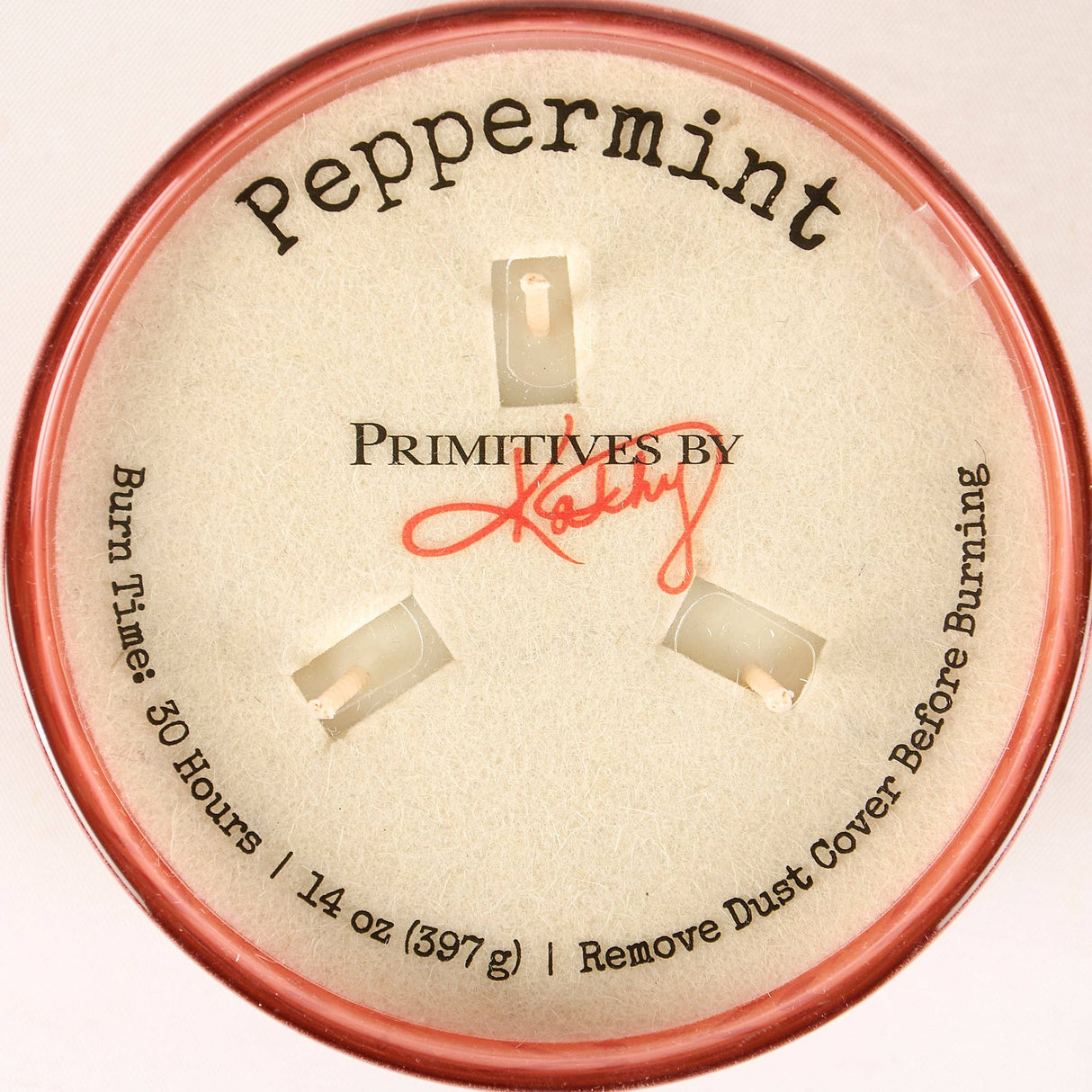 A round tin of Primitives by Kathy Peppermint scented burns, showing the beige inner lid with the brand name, scent 'Peppermint', and burn time details, framed by a pink-colored rim.
