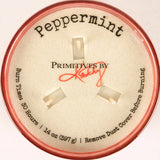 A round tin of Primitives by Kathy Peppermint scented burns, showing the beige inner lid with the brand name, scent 'Peppermint', and burn time details, framed by a pink-colored rim.