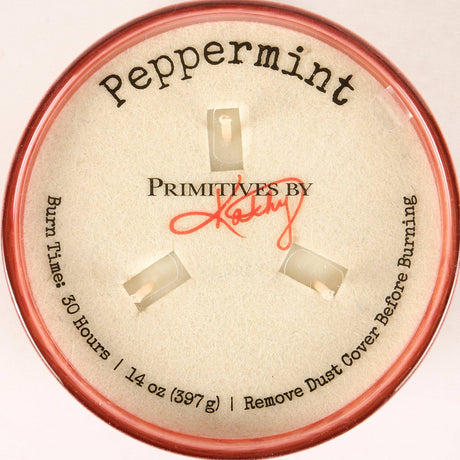 A round tin of Primitives by Kathy Peppermint scented burns, showing the beige inner lid with the brand name, scent 'Peppermint', and burn time details, framed by a pink-colored rim.