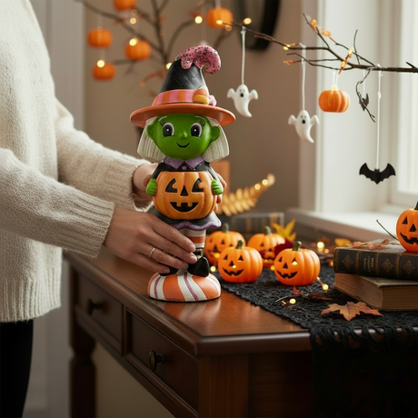 Person holding a colorful Halloween witch figurine in a festive display with pumpkins, fall décor, black gauze cloth, and hanging ghost and pumpkin accents.