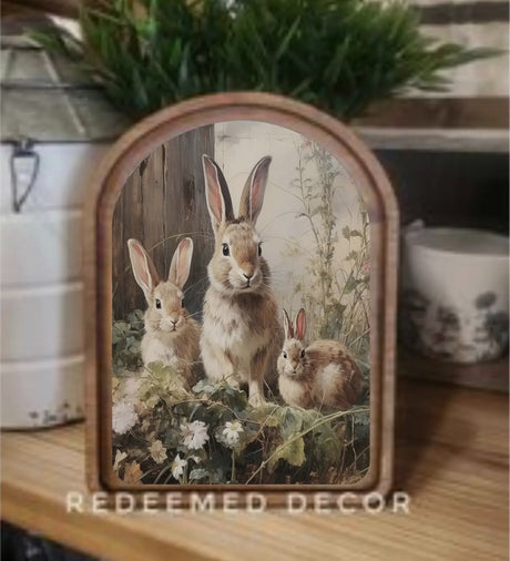 Three rabbits in a spring meadow with wildflowers, featured in an arched-top print from Redeemed Decor.
