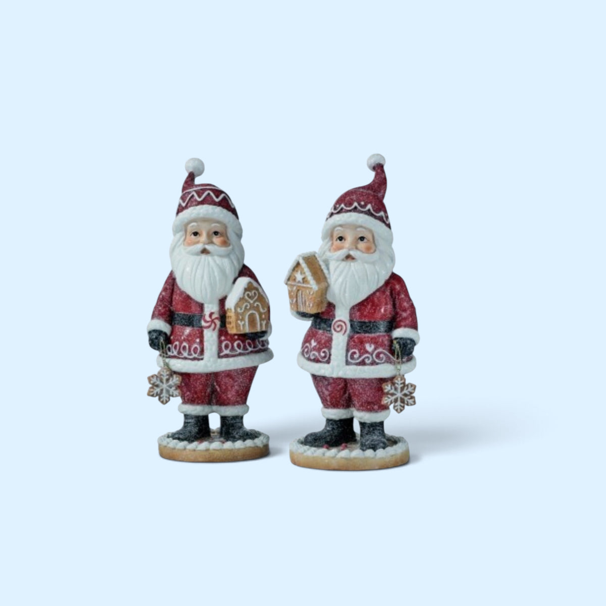 Two glittered Santa figurines stand on icing-inspired cookie bases while holding decorated gingerbread houses. One Santa holds a gingerbread house with a heart detail, while the other holds a gingerbread house featuring a star detail. Each Santa wears a red glitter coat accented with white icing-inspired trim and carries a white snowflake ornament.