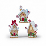 Three illuminated gingerbread-style houses feature snowy white icing details, candy cane accents, and warm glowing windows. The collection includes a Snowflake House with a snowflake chimney topper, a Peppermint House with peppermint candy accents, and a Bow House adorned with a large red bow. Each resin cottage is illuminated from within for a cozy holiday glow.