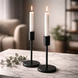 Two white taper candles in sleek black modern holders on a marble surface with soft, cozy background lighting.