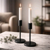 Two white taper candles in sleek black modern holders on a marble surface with soft, cozy background lighting.