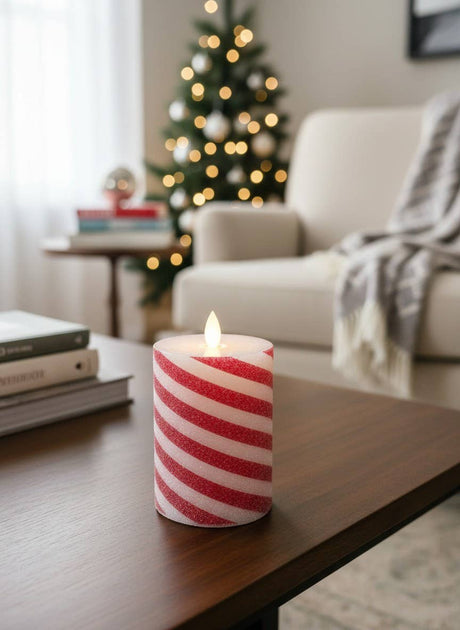Red‑and‑white candy cane striped LED pillar candle glowing on a wooden coffee table in a cozy holiday living room, with a softly lit Christmas tree and a blanket‑draped armchair in the background.