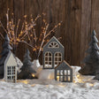 Cozy winter village display with miniature houses, evergreen trees, warm string lights, and a soft layer of artificial snow creating a festive holiday atmosphere.