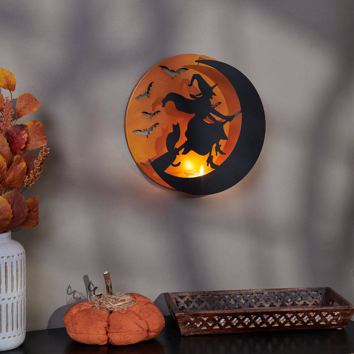 Metal wall candle holder featuring a witch flying across the moon with bat accents, illuminated by a tealight for warm Halloween ambiance.