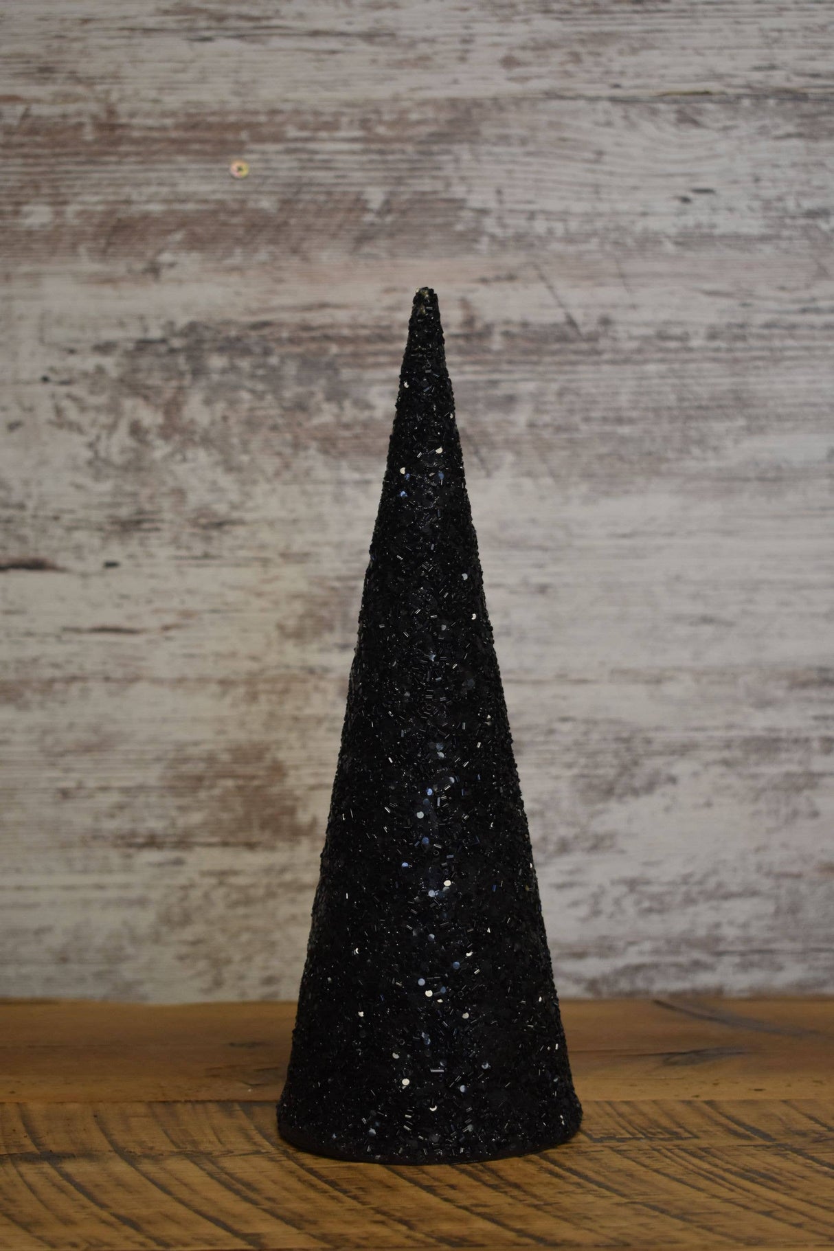 Black glass‑beaded cone tree displayed on a wooden surface with a rustic backdrop, showcasing its sleek shape and shimmering texture.
